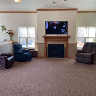 A cozy living room with a television mounted above a fireplace, surrounded by comfortable armchairs and windows letting in natural light.
