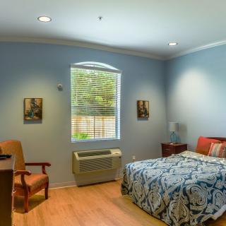A cozy and well-lit bedroom featuring a bed with patterned bedding, a comfortable chair, and an air conditioning unit beneath a window.