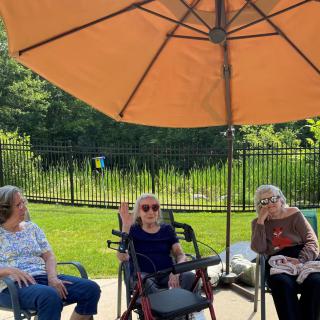 A group of seniors sitting under a large umbrella in a garden on a sunny day.