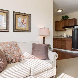 A cozy living room with a sofa and decorative pillows adjacent to a kitchen area, featuring warm lighting and artwork on the walls.