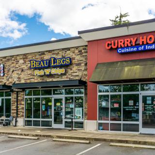 A view of a small shopping center featuring 'Beau Legs Fish & Chips' and 'Curry House Cuisine of India' with a parking area in front.
