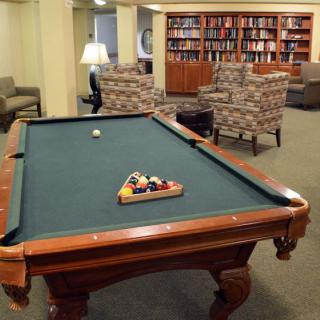 A cozy library room featuring a pool table set with balls, surrounded by bookshelves and comfortable seating.