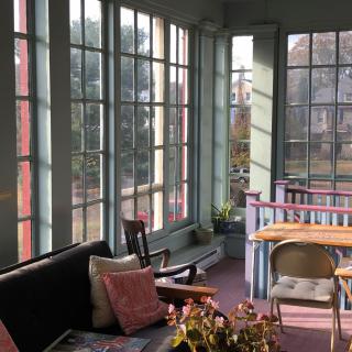 A cozy sunroom filled with natural light, featuring a sofa, chairs, and a table surrounded by large windows.