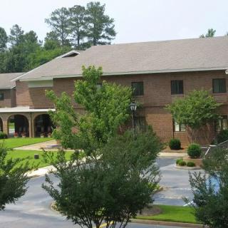 A two-story brick building surrounded by trees with a parking lot in the foreground.