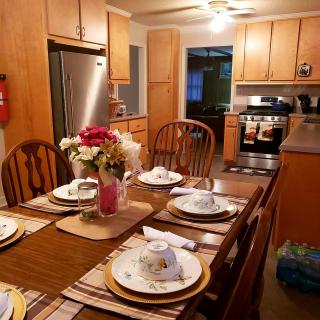 A cozy kitchen with a wooden dining table set for six, featuring floral decorations and surrounded by wooden cabinets and modern appliances.
