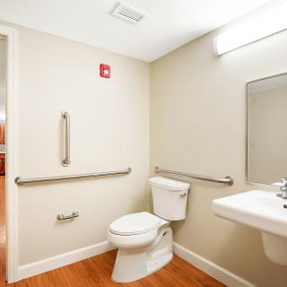 An accessible bathroom with grab bars, featuring a toilet, sink, and mirror on a wooden floor.