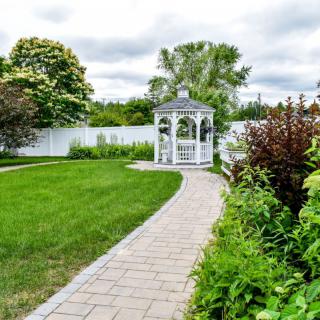 A beautiful garden with a brick pathway leading to a white gazebo surrounded by lush greenery.