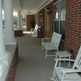 A view of a porch with white rocking chairs and columns along a brick building.