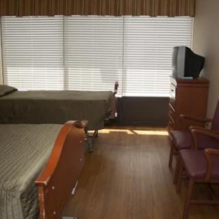 A hospital room with two beds, a television, and chairs in front of large windows with blinds.