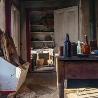 An abandoned room with a cluttered table and shelves filled with various items, illuminated by natural light.