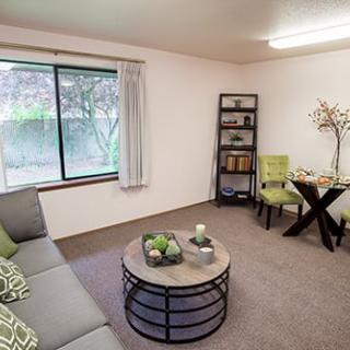 A cozy living room with modern furnishings, featuring a sofa, coffee table, bookshelf, and window view.