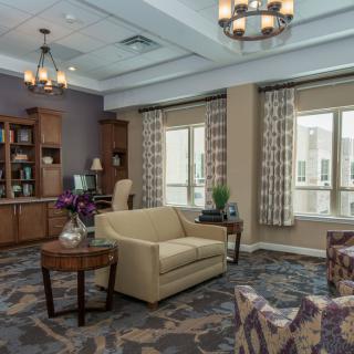 A cozy living room with elegant furniture and decor, featuring a sofa, armchair, coffee table, bookshelves, and large windows with curtains.