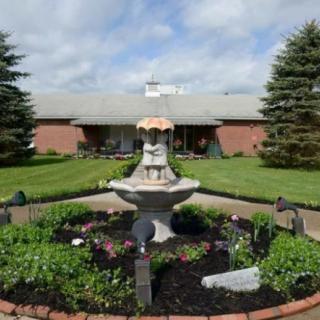 A garden with a central fountain surrounded by flowers and greenery, in front of a brick building.