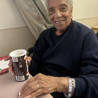 An elderly person smiling while holding a cup indoors.