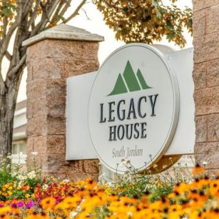 A sign for Legacy House amidst colorful flowers and a brick wall backdrop.