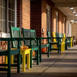 A row of colorful benches and chairs with pumpkins outside a brick building.