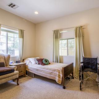 A simple bedroom with a hospital bed, wheelchair, and armchair, featuring natural light from two windows.