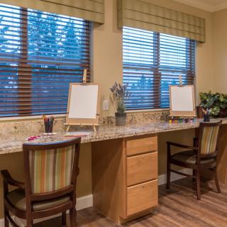 A cozy office workspace with wooden desks and chairs, featuring large windows with blinds.