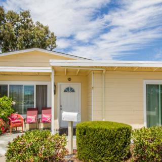 A cozy yellow house with a small front porch featuring red chairs, surrounded by neatly trimmed bushes under a partly cloudy sky.