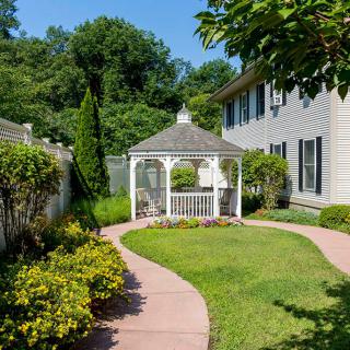 A well-maintained garden featuring a gazebo, surrounded by lush greenery and a paved pathway leading to a house.