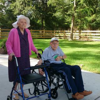 An elderly couple enjoying a sunny day outdoors, with one using a walker and the other in a wheelchair.