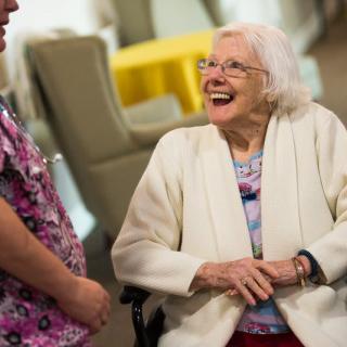 An elderly woman in a wheelchair is laughing and conversing with a caregiver indoors.