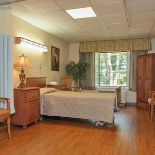 A hospital room with a single bed, wooden furniture, and a large window offering natural light.