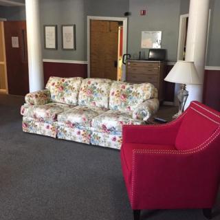 A cozy lobby area featuring a floral-patterned sofa, a red armchair, and a lamp.