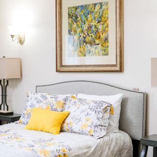 A well-decorated bedroom featuring a bed with floral bedding, a yellow accent pillow, stylish lamps, and framed artwork.