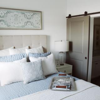 A minimalist bedroom with a neatly made bed, featuring blue and white pillows and a decorative wall piece. A lamp sits on a side table, and a sliding door is visible in the background.