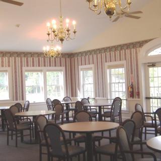An empty dining room with several round tables and chairs, decorated with chandeliers and lined with windows and curtains.