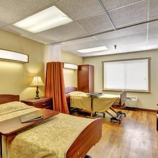 A well-lit hospital room with two beds separated by a privacy curtain and equipped with bedside tables and lamps.
