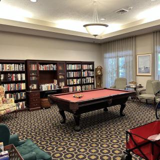 A cozy library room features bookshelves filled with books and a billiards table at the center, surrounded by comfortable chairs.