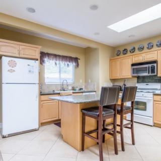 A modern kitchen interior with wooden cabinetry, featuring a fridge, stove, microwave, and a central island with bar stools.