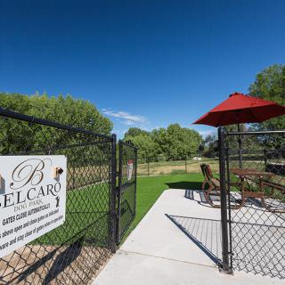 A fenced dog park with a red umbrella and seating area on a sunny day.