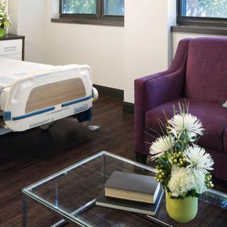 A well-furnished hospital room with a bed, a purple chair, and a table holding flowers.