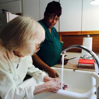 An elderly woman receives assistance while washing her hands in the kitchen.