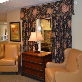 A cozy living room setup featuring a pair of beige armchairs, a wooden dresser with a lamp, and floral curtains.