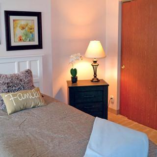 A cozy bedroom with a neatly made bed, featuring a decorative pillow, a nightstand with a lamp and plant, and framed artwork on the wall.