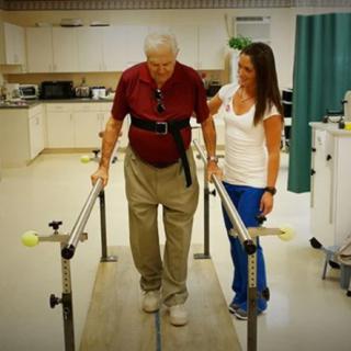 An elderly man receiving assistance from a therapist during physical rehabilitation.