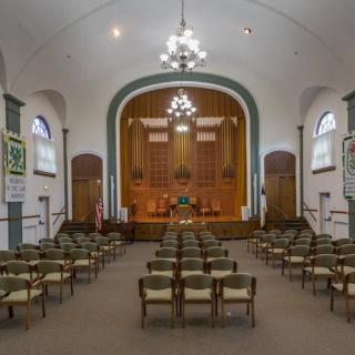 Interior view of a church with rows of chairs facing a raised stage area, featuring chandeliers and arched windows.