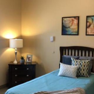 A cozy bedroom with a single bed, decorated with colorful pillows and paintings, alongside a nightstand and lamp.