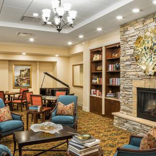 A cozy lounge featuring a stone fireplace, comfortable seating, bookshelves, and a piano.