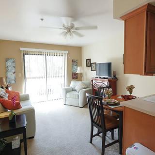 A cozy living room with a sofa, television, and dining area, featuring warm decor and natural light from large windows.