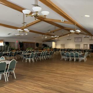 An empty banquet hall with arranged tables and chairs, featuring a wooden floor and ceiling lights.
