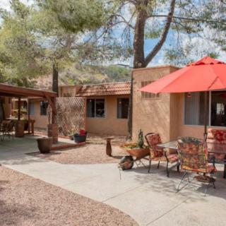 A serene courtyard patio with outdoor furniture and a red umbrella, surrounded by trees and an adobe-style house.