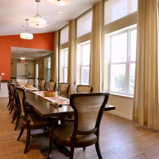A spacious room featuring a long table with chairs, large windows with curtains, and elegant lighting.