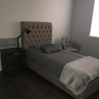 A minimalist bedroom featuring a single bed with gray bedding, a nightstand with a lamp, and an accent chair.