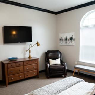 A cozy bedroom with a TV mounted on the wall, a wooden dresser, an armchair, and a large window.