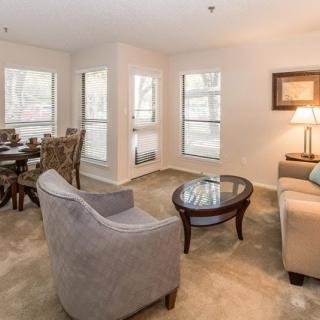A cozy living room with a sofa and armchair, complemented by a dining area and ample natural light from multiple windows.